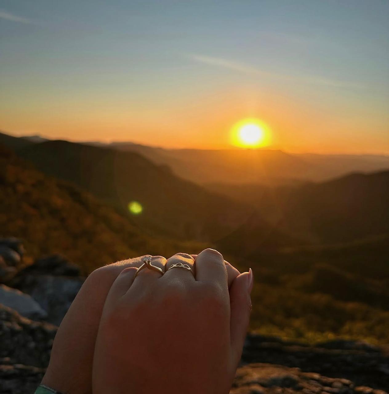 Pôr do sol dourado com as mãos de Thaynara e Pedro entrelaçadas, exibindo as alianças de noivado.